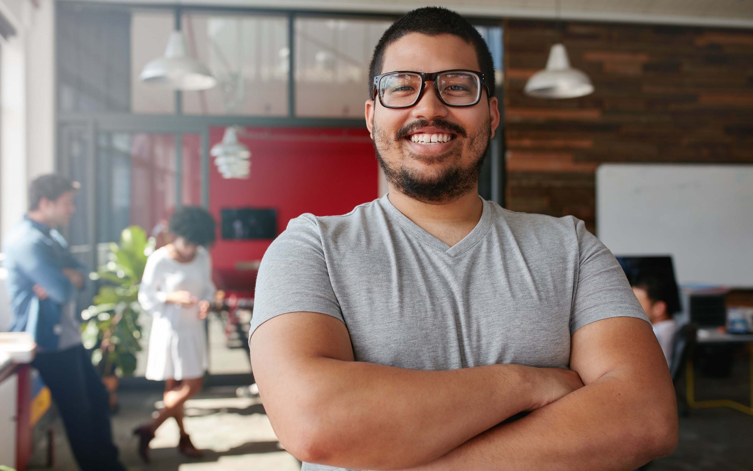Portrait of a confident creative professional with colleagues in the background. Mixed race man standing with his arms crossed looking at camera and smiling.